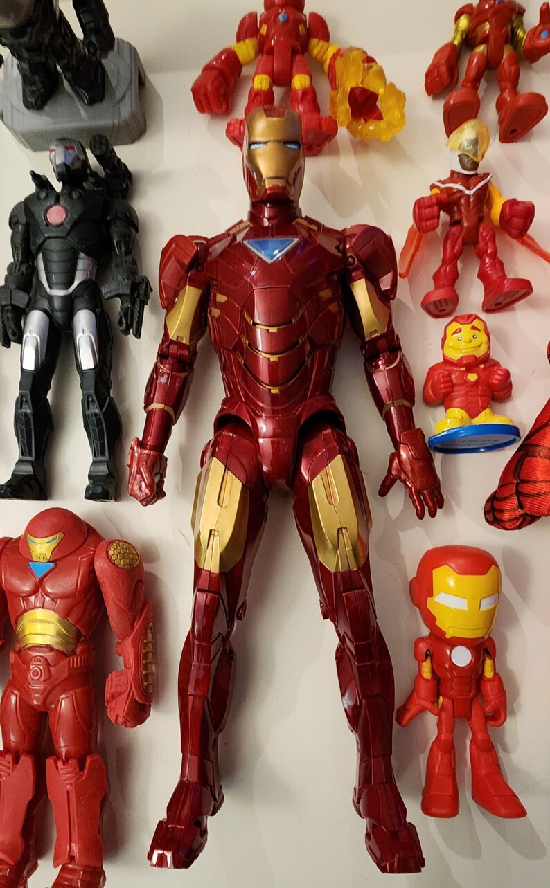 May include: A collection of Iron Man action figures in various sizes and styles. The figures are primarily red and gold, with some featuring black and silver accents. The central figure is a large, detailed Iron Man with a metallic finish.