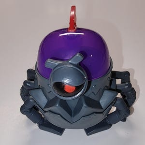 May include: A toy robot with a purple dome head and grey body. The robot has a single red eye and multiple articulated limbs. A red loop extends from the top of the head. The robot is designed with a futuristic aesthetic.