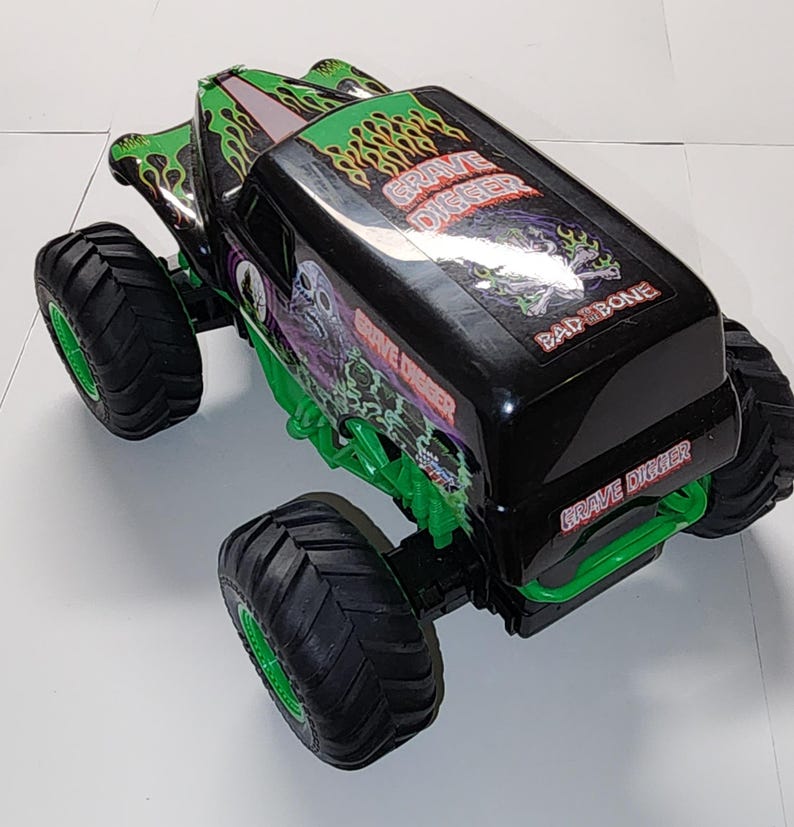 Puede incluir: Juguete de cami&oacute;n monstruo Grave Digger negro y verde con neum&aacute;ticos negros grandes y llantas verdes. El cami&oacute;n presenta el logotipo de Grave Digger y gr&aacute;ficos de llamas. El juguete tiene un dise&ntilde;o Bad Bone en la parte superior.