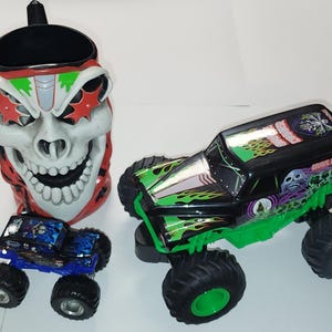 Puede incluir: Colecci&oacute;n de art&iacute;culos con tem&aacute;tica de monster trucks. Incluye una taza con forma de calavera con detalles rojos y blancos, y dos monster trucks de juguete, uno azul y otro negro y verde, con neum&aacute;ticos grandes.