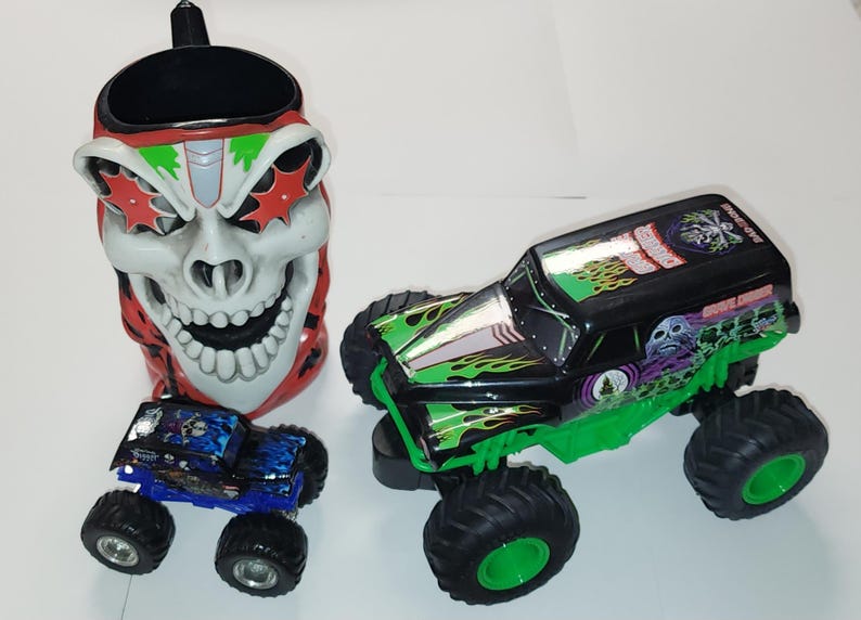 Puede incluir: Colecci&oacute;n de art&iacute;culos con tem&aacute;tica de monster trucks. Incluye una taza con forma de calavera con detalles rojos y blancos, y dos monster trucks de juguete, uno azul y otro negro y verde, con neum&aacute;ticos grandes.