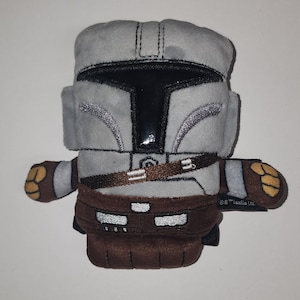May include: A plush toy depicting a Mandalorian helmet and armor. The toy is primarily gray with black and brown accents. The helmet has a shiny black visor and silver detailing. The toy has brown gloves and boots, and a brown belt.
