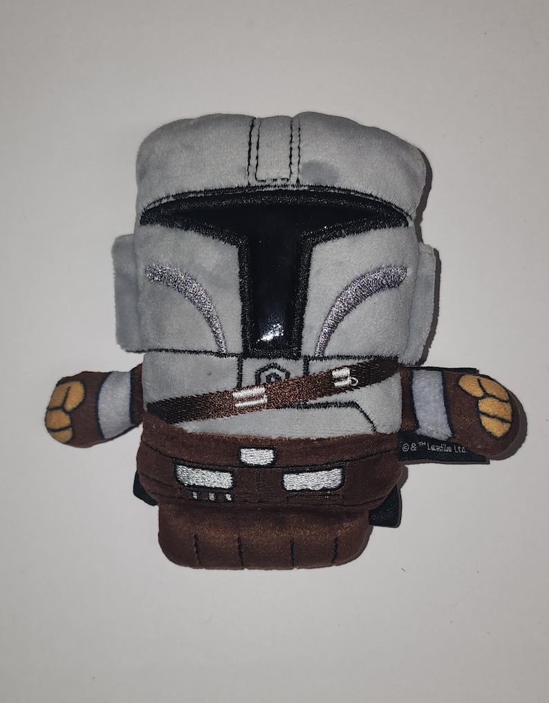 May include: A plush toy depicting a Mandalorian helmet and armor. The toy is primarily gray with black and brown accents. The helmet has a shiny black visor and silver detailing. The toy has brown gloves and boots, and a brown belt.