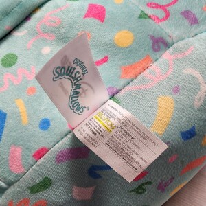 Pode incluir: Um close-up de um brinquedo de pel&uacute;cia com um fundo azul claro coberto por formas de confetes coloridos. A etiqueta diz "Original Squishmallows" e inclui as informa&ccedil;&otilde;es do distribuidor. O brinquedo &eacute; feito de fibras de poli&eacute;ster e feltro.