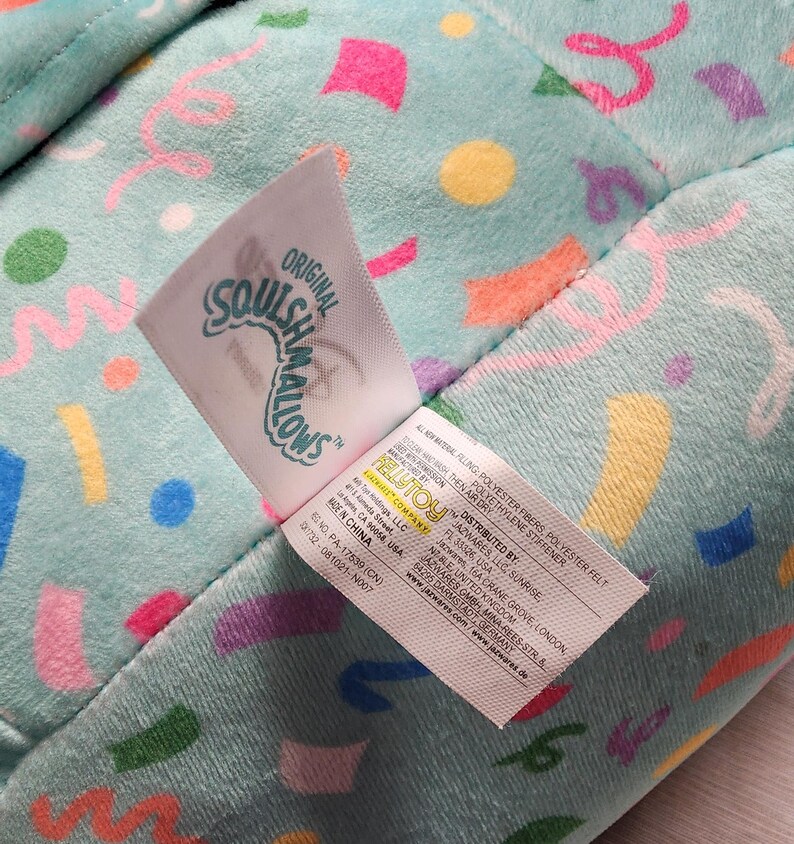 Pode incluir: Um close-up de um brinquedo de pel&uacute;cia com um fundo azul claro coberto por formas de confetes coloridos. A etiqueta diz "Original Squishmallows" e inclui as informa&ccedil;&otilde;es do distribuidor. O brinquedo &eacute; feito de fibras de poli&eacute;ster e feltro.