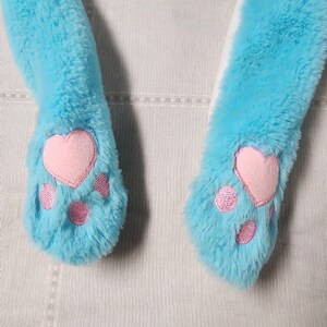 May include: Close-up of a pair of light blue plush cat paw mittens. Each mitten features a pink heart-shaped paw pad and three smaller pink paw pads. The mittens are made of soft, fluffy material.