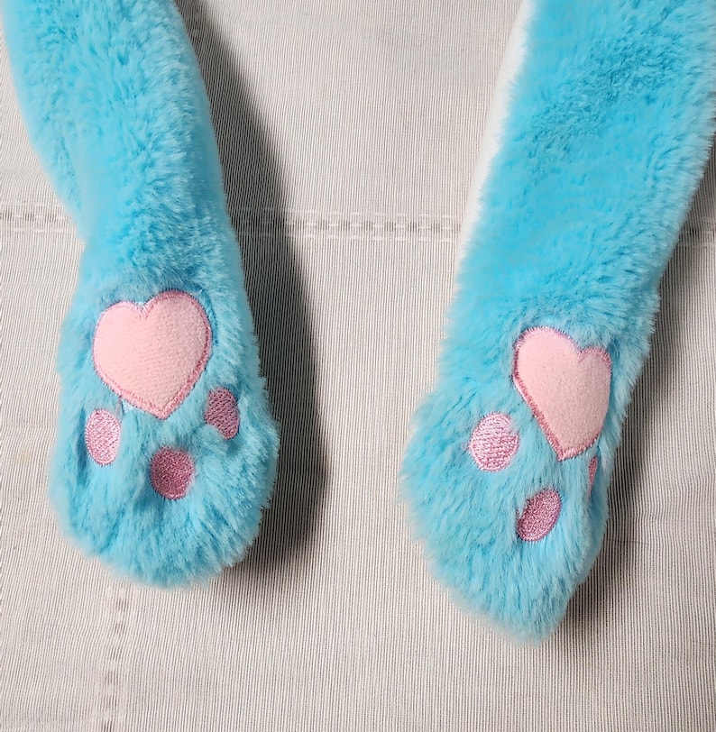 May include: Close-up of a pair of light blue plush cat paw mittens. Each mitten features a pink heart-shaped paw pad and three smaller pink paw pads. The mittens are made of soft, fluffy material.