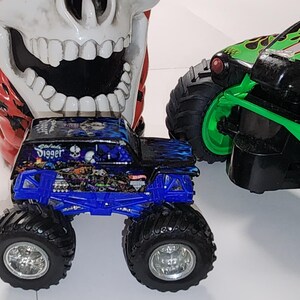 Puede incluir: Un juguete de cami&oacute;n monstruo azul y negro con la palabra "Digger" en el lateral, junto a un juguete de cami&oacute;n monstruo verde y negro. Un gran recipiente con forma de calavera est&aacute; en el fondo.