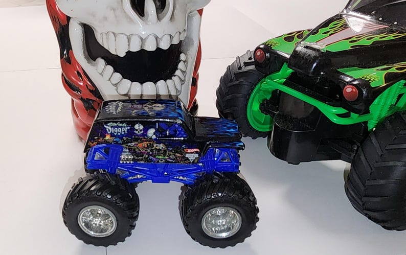 Puede incluir: Un juguete de cami&oacute;n monstruo azul y negro con la palabra "Digger" en el lateral, junto a un juguete de cami&oacute;n monstruo verde y negro. Un gran recipiente con forma de calavera est&aacute; en el fondo.