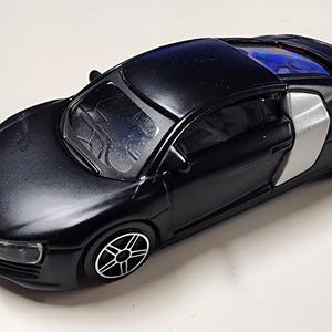 Puede incluir: Un coche de juguete negro con detalles plateados. El coche de juguete presenta un diseño elegante, ruedas detalladas y el logotipo de Audi en la parte delantera. El coche mide aproximadamente 13 cm de largo.