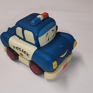Puede incluir: Un coche de policía de juguete azul y blanco con una cara sonriente. El coche tiene una barra de luces roja y azul en la parte superior, la palabra "POLICE" en el lateral y ruedas rosas. El coche es un diseño de dibujos animados.