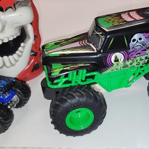 Puede incluir: Imagen de dos camiones monstruo de juguete y una m&aacute;scara de calavera. El cami&oacute;n m&aacute;s grande es negro y verde con gr&aacute;ficos de "Grave Digger". El cami&oacute;n m&aacute;s peque&ntilde;o es azul. La m&aacute;scara de calavera es blanca con detalles rojos.