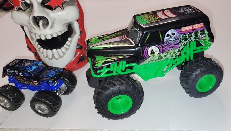 Puede incluir: Imagen de dos camiones monstruo de juguete y una m&aacute;scara de calavera. El cami&oacute;n m&aacute;s grande es negro y verde con gr&aacute;ficos de "Grave Digger". El cami&oacute;n m&aacute;s peque&ntilde;o es azul. La m&aacute;scara de calavera es blanca con detalles rojos.