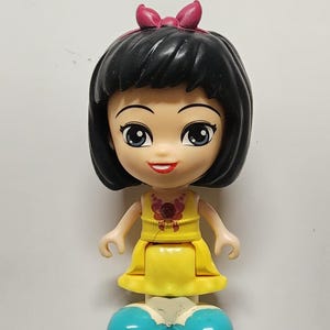 May include: A plastic doll with black hair, a pink bow, and a yellow dress. The doll has blue shoes and a necklace. The doll is smiling and has large eyes. The doll is a toy.