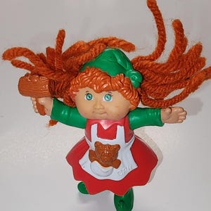 May include: A vintage Cabbage Patch Kids figurine of a girl with orange yarn hair, blue eyes, and a green cap and sleeves. She wears a red dress and a white apron with a teddy bear design. The figurine is holding a brown object.