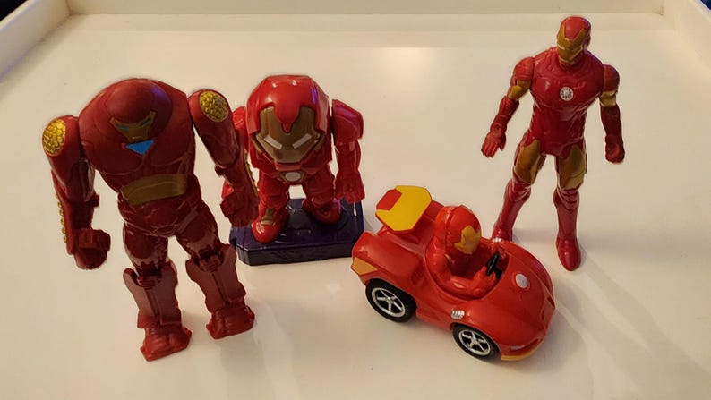 May include: A collection of red and gold Iron Man toys, including two action figures, a small car, and a larger robot-like figure. The toys are made of plastic and are set against a white background.