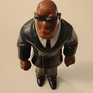 May include: A figurine of a bald, dark-skinned man wearing a black suit, tie, and sunglasses. The figure has a stern expression and is posed in a standing position. The suit has a white collared shirt underneath. The figure is a collectible.