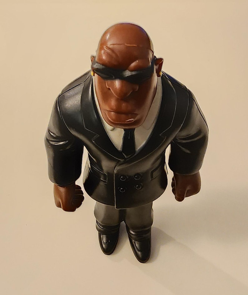 May include: A figurine of a bald, dark-skinned man wearing a black suit, tie, and sunglasses. The figure has a stern expression and is posed in a standing position. The suit has a white collared shirt underneath. The figure is a collectible.