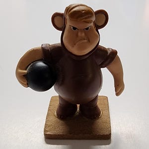 May include: A brown figurine of a cartoon character with an angry expression, wearing a brown outfit with monkey ears and holding a black ball. The figurine is standing on a small, square, wooden base.