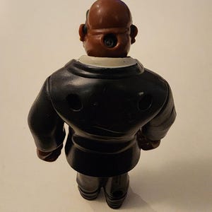 May include: A vintage toy figurine of a man in a black suit, viewed from the back. The figure has a brown head and a white collar. The suit has two circular holes. The toy is standing on a light-colored surface.