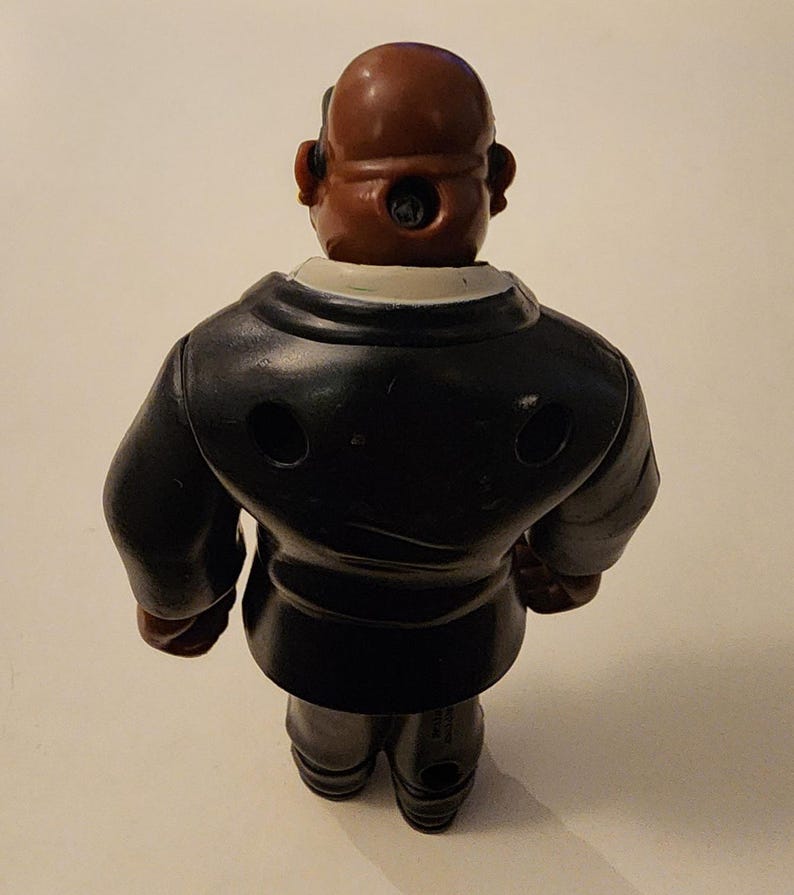 May include: A vintage toy figurine of a man in a black suit, viewed from the back. The figure has a brown head and a white collar. The suit has two circular holes. The toy is standing on a light-colored surface.