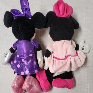 May include: Two plush Minnie Mouse dolls. One doll wears a purple dress with pink polka dots and a matching bow. The other doll is dressed in a pink dress and a pink hat. Both dolls have black ears and black shoes.