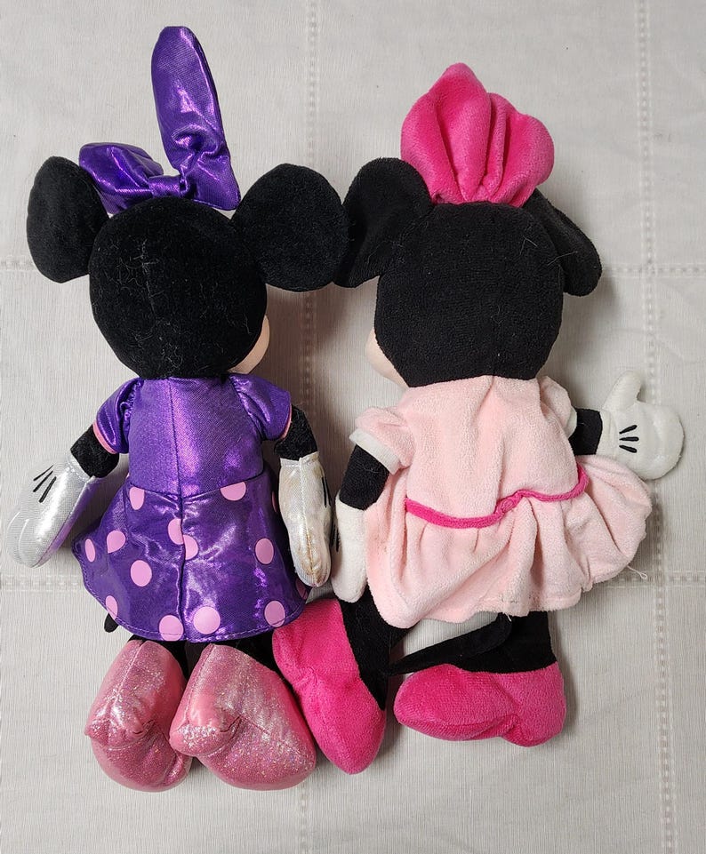 May include: Two plush Minnie Mouse dolls. One doll wears a purple dress with pink polka dots and a matching bow. The other doll is dressed in a pink dress and a pink hat. Both dolls have black ears and black shoes.