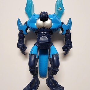 Op de afbeelding: Een blauw en marineblauw actiefiguur met een gestileerd insectachtig ontwerp. De figuur heeft grote, vleugelachtige structuren, zilveren borstdetails en beweegbare ledematen. Het plastic speelgoed heeft een gedetailleerd, getextureerd oppervlak.