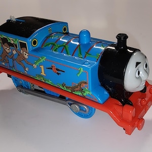 以下が含まれることがあります： 黒い屋根と赤いアクセントが付いた青いおもちゃの電車。電車には笑顔の顔があり、猿や鳥など、ジャングルをテーマにしたグラフィックで装飾されています。番号1が側面に表示されています。