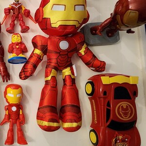 May include: A collection of Iron Man toys, including a plush doll, a small figurine, and a red car with yellow accents. The plush doll is red and yellow, with a large head and a round white circle on the chest. The car has the words "Iron Man" and "Armored Avenger" printed on it.