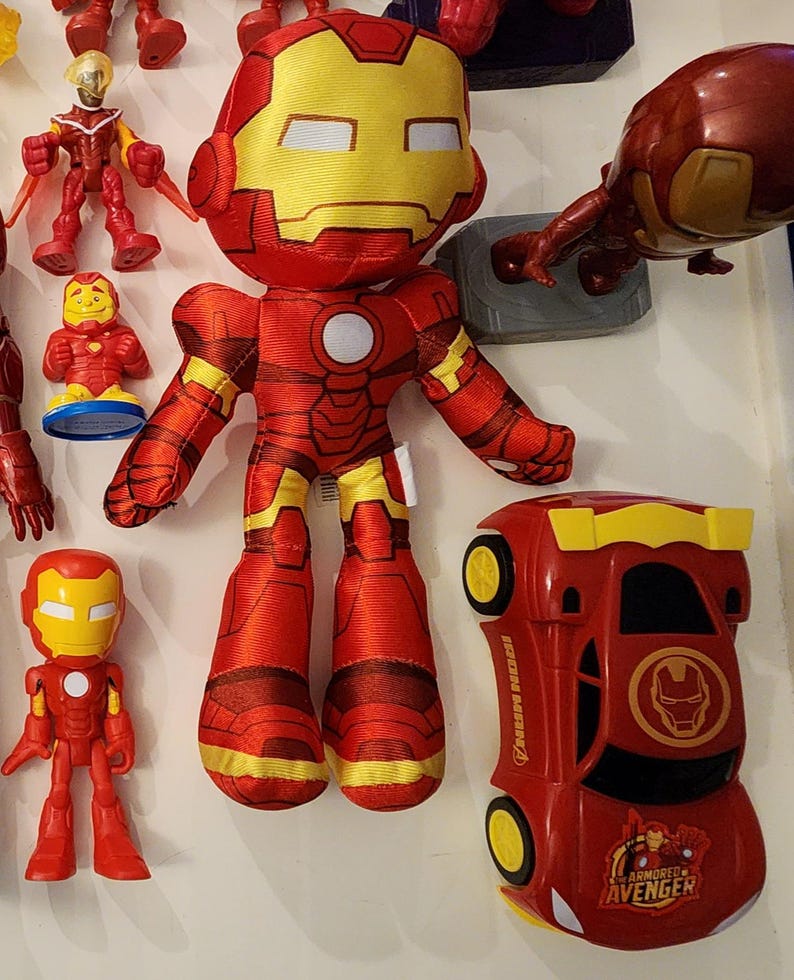 May include: A collection of Iron Man toys, including a plush doll, a small figurine, and a red car with yellow accents. The plush doll is red and yellow, with a large head and a round white circle on the chest. The car has the words "Iron Man" and "Armored Avenger" printed on it.