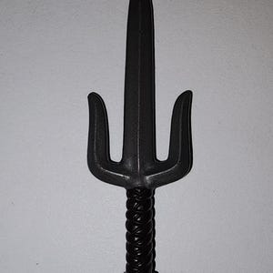 Puede incluir: Un sai negro, un arma tradicional japonesa, se muestra sobre un fondo blanco. El sai presenta una hoja central con dos púas curvas que se extienden desde el mango. El mango está envuelto con un diseño texturizado en forma de cuerda.