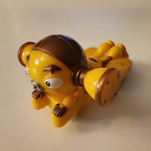 Könnte beinhalten: Eine gelbe Spielfigur mit braunem Helm und großen, runden Ohrenteilen. Die Figur hat ein lächelndes Gesicht mit großen Augen und kleinen Händen. Das Spielzeug liegt auf dem Rücken.