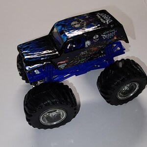 Puede incluir: Un juguete de cami&oacute;n monstruo negro y azul con neum&aacute;ticos negros grandes. El cami&oacute;n tiene un dise&ntilde;o de llama azul en el cap&oacute; y las palabras "Grave Digger" en el techo. El chasis es azul.