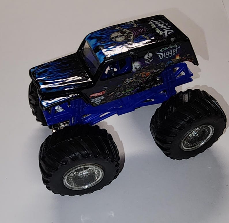 Puede incluir: Un juguete de cami&oacute;n monstruo negro y azul con neum&aacute;ticos negros grandes. El cami&oacute;n tiene un dise&ntilde;o de llama azul en el cap&oacute; y las palabras "Grave Digger" en el techo. El chasis es azul.