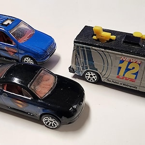 May include: Three toy cars: a blue SUV, a black coupe with Superman logos, and a gray news van with "News 12 Live" text. The vehicles are die-cast models with detailed features and rolling wheels.