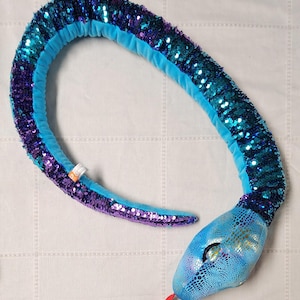 May include: A plush snake toy in shades of blue and purple. The body is covered in sequins, with a light blue underside. The head is a lighter blue with a textured pattern and a red tongue. A fun and colorful toy.