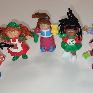Op de afbeelding: Een verzameling van zes vintage Cabbage Patch Kids poppen. De poppen dragen verschillende outfits, waaronder een rood en blauw militair uniform, een groene en rode jurk en een paarse jurk. Elke pop heeft een unieke haarstijl.
