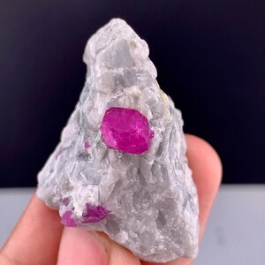 May include: A raw mineral specimen featuring a jagged, light gray, crystalline matrix embedded with several vibrant, deep pink ruby crystals. The stone has a rough, uneven texture with prominent facets and natural inclusions throughout the structure.