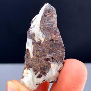 May include: A raw mineral specimen with a mix of brown, white, and tan colors. The rough, irregular shape of the stone is held by a person's fingers, showcasing its natural texture and crystalline structure. The background is a dark, solid color.