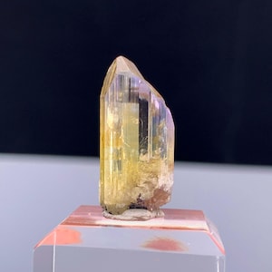 May include: A translucent, yellow-orange crystal with a pointed top and a rough base. The crystal has vertical striations and a hint of purple on one side. It is displayed on a clear, rectangular stand with a pinkish-orange top.