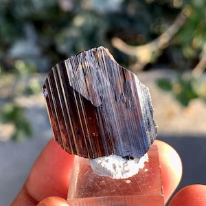 Puede incluir: Un espécimen mineral oscuro y metálico con una apariencia en capas y estriada. El mineral es de color marrón oscuro con toques de púrpura y se asienta sobre una base rectangular transparente con un material blanco texturizado.