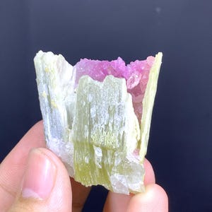 May include: A raw, natural mineral specimen featuring a combination of colors. The outer layer is a pale green, with a vibrant pink interior. The crystal structure is visible, with a rough, textured surface. The mineral is held in a hand.
