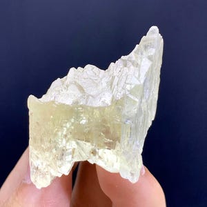 Könnte beinhalten: Eine durchscheinende, hellgelbe Kristallformation mit einer gezackten, strukturierten Oberfläche. Der Kristall hat ein geschichtetes Aussehen und wird vor einem dunkelblauen Hintergrund gehalten. Die Gesamtform ist unregelmäßig und natürlich.