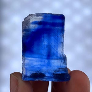 May include: A vibrant blue crystal specimen with a rectangular shape. The crystal has a translucent quality, with varying shades of blue and white. The rough edges and natural texture of the crystal are visible.