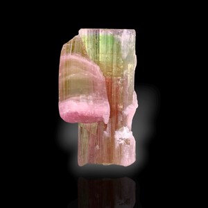 May include: A raw tourmaline crystal specimen featuring a vertical columnar structure with a distinct colour gradient transitioning from deep pink at the base and side to a translucent light green at the top, displaying natural striations and textures.