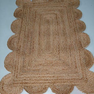 Könnte beinhalten: Ein rechteckiger Jute-Teppich mit einem gewellten Rand. Der Teppich hat eine natürliche beige Farbe und eine gewebte Textur. Der gewellte Rand verleiht dem Design des Teppichs eine dekorative Note. Dieser Teppich eignet sich für verschiedene Einrichtungsstile.