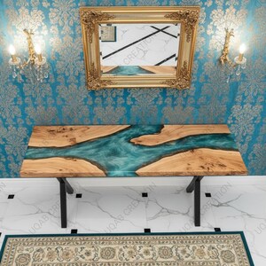 May include: A wooden and teal epoxy resin table with black metal legs. The table is rectangular and has a river-like design. A gold-framed mirror hangs above the table, reflecting the table's design. The background features a blue wallpaper with a gold floral pattern.