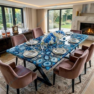 Puede incluir: Una mesa de comedor rectangular con una llamativa tapa de piedra de ágata azul. La mesa está puesta con platos, cubiertos y cristalería. Seis sillas de terciopelo marrón rodean la mesa. Un arreglo floral y una chimenea están en el fondo.