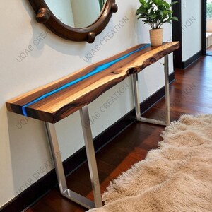 May include: A live edge wooden console table with a blue resin river. The table has a natural wood grain finish and sits on silver metal legs. A small potted plant sits on the table's surface. The table is approximately 60 inches long.